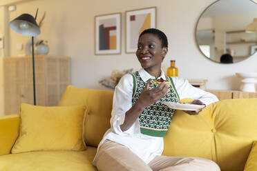 Smiling woman having breakfast on yellow sofa at home - JCZF00883