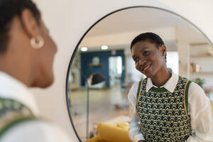 Lächelnde Frau betrachtet ihr Spiegelbild in der Wohnung stehend - JCZF00886