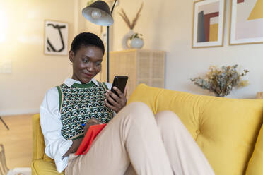 Lächelnde Frau, die auf dem Sofa im Wohnzimmer sitzt und auf ihr Mobiltelefon schaut - JCZF00900