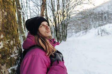 Contemplative man in knit hat leaning on tree in snowy forest - OMIF00377