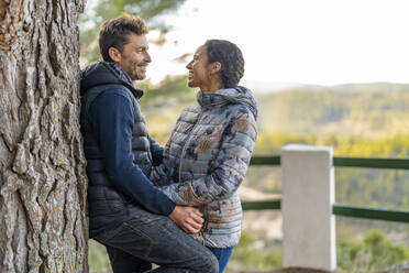 Happy couple looking at each other standing by tree trunk - DLTSF02539