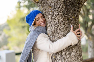 Happy woman with eyes closed embracing tree trunk - DLTSF02547