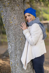 Lächelnde Frau lehnt an einem Baumstamm im Park - DLTSF02548