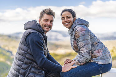 Smiling couple wearing padded jackets sitting at park - DLTSF02551