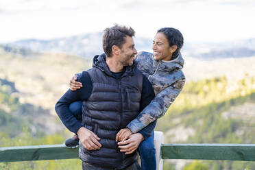 Smiling woman sitting on railing hugging man from behind at park - DLTSF02552