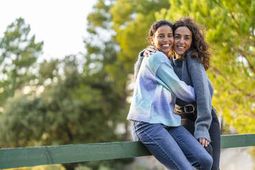 Multiracial friends embracing each other sitting on railing at park - DLTSF02572