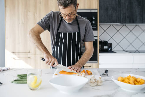 Mann mit Brille schneidet Karotte auf Kücheninsel zu Hause - JRFF05225