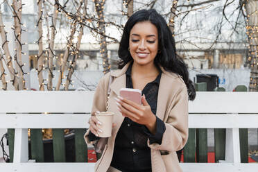 Lächelnde Frau mit Kaffeetasse und Smartphone auf einer Bank - JRVF02396