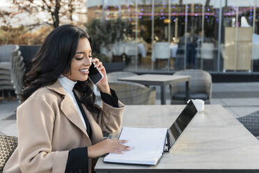 Geschäftsfrau mit Tagebuch und Laptop, die in einem Café mit ihrem Mobiltelefon spricht - JRVF02420