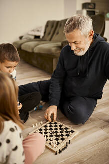 Smiling grandfather playing chess with grandchildren at home - ZEDF04360