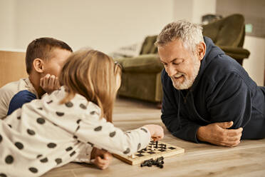 Grandfather looking at girl playing chess lying on floor at home - ZEDF04366
