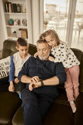 Boy and girl sitting with grandfather using smart watch sitting on sofa at home - ZEDF04381