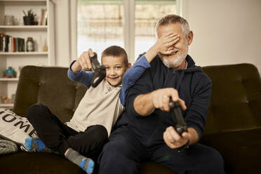 Grandson covering eyes of grandfather playing video game at home - ZEDF04393