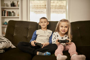 Brother and sister playing video game sitting on sofa at home - ZEDF04396