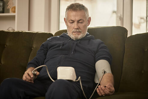 Älterer Mann beim Blutdruckmessen auf dem Sofa zu Hause - ZEDF04410