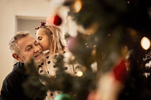 Granddaughter kissing grandfather behind christmas tree at home - ZEDF04419