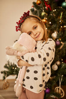 Nettes Mädchen umarmt Plüschtier vor dem Weihnachtsbaum zu Hause stehen - ZEDF04423