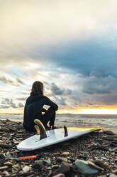 Surfer sitzt mit Surfbrett auf Kieselsteinen und betrachtet den Sonnenuntergang über dem Meer - OMIF00422