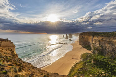 Australien, Victoria, Blick auf die Sonne, die durch die Sturmwolken über dem Sandstrand im Port Campbell National Park scheint, mit den Zwölf Aposteln im Hintergrund - FOF12591