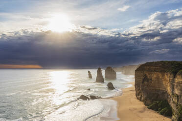 Australien, Victoria, Blick auf die Sonne, die durch die Sturmwolken über dem Sandstrand im Port Campbell National Park scheint, mit den Zwölf Aposteln im Hintergrund - FOF12593