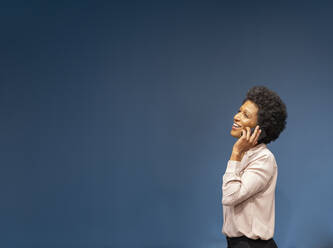 Happy businesswoman talking on smart phone by blue wall in office - EIF03038