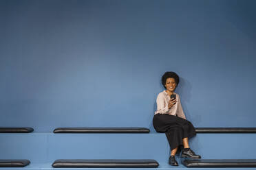 Smiling businesswoman using mobile phone in front of blue wall at office - EIF03041