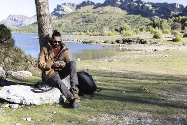 Wanderer mit Mobiltelefon auf einem Felsen sitzend am Cuber-Stausee, Mallorca, Balearen, Spanien - JPTF01019