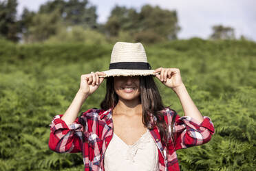 Smiling woman wearing hat in forest on sunny day - WPEF05645