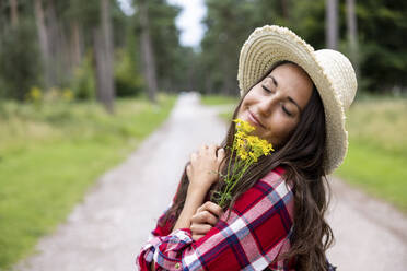 Smiling woman with hat holding yellow daisy flowers on road at Cannock Chase - WPEF05676