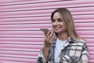 Young woman talking on speaker phone near pink shutter - PNAF02772