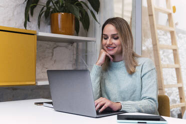 Lächelnde Geschäftsfrau mit Hand am Kinn bei der Arbeit am Laptop im Studio - PNAF02773