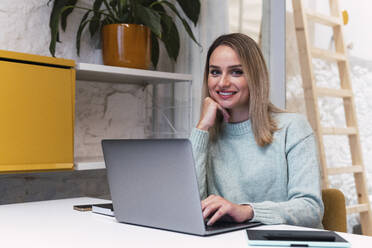 Beautiful businesswoman with hand on chin sitting with laptop at studio - PNAF02774