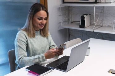 Smiling businesswoman using smart phone at desk in studio - PNAF02779