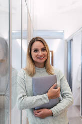 Lächelnde Geschäftsfrau mit Laptop, die sich in einem Studio an ein Glas lehnt - PNAF02795