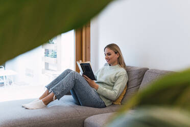 Woman reading book sitting on sofa at home - PNAF02813