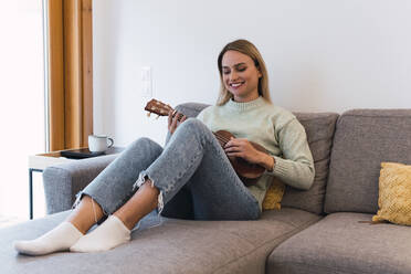 Smiling woman playing ukulele sitting on sofa at home - PNAF02814