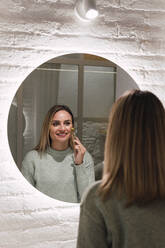 Smiling woman using jade roller looking in mirror on wall at home - PNAF02818