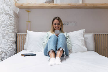Frau mit Smartphone auf dem Bett sitzend zu Hause - PNAF02823