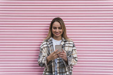 Frau, die ein Mobiltelefon vor einem rosa Fensterladen benutzt - PNAF02826