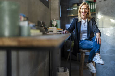 Smiling businesswoman sitting on stool by desk in office - DLTSF02588