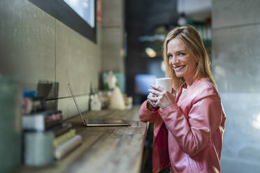 Smiling businesswoman with laptop on desk holding coffee mug at office - DLTSF02601