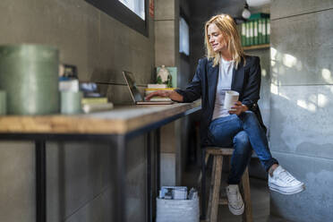 Geschäftsfrau mit Kaffeebecher und Laptop auf einem Hocker im Büro sitzend - DLTSF02604