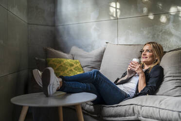 Businesswoman holding coffee mug sitting on sofa in office - DLTSF02606