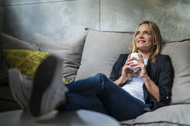 Smiling businesswoman holding coffee mug day dreaming on sofa in office - DLTSF02607