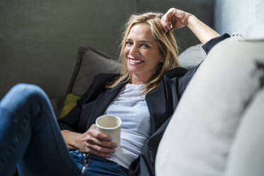 Smiling businesswoman holding coffee mug sitting on sofa - DLTSF02608