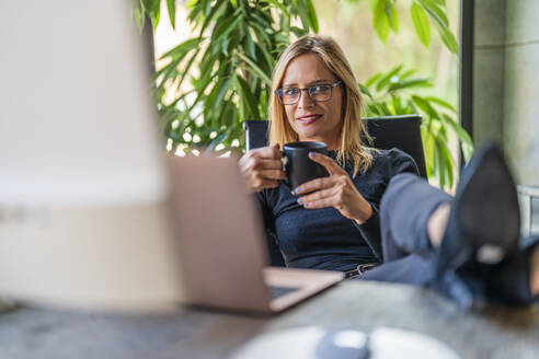 Geschäftsfrau mit Kaffeetasse, die mit hochgelegten Füßen am Schreibtisch sitzt - DLTSF02616