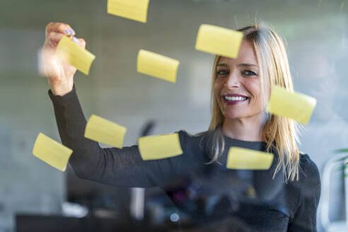 Smiling businesswoman writing on adhesive notes seen through glass wall at office - DLTSF02622