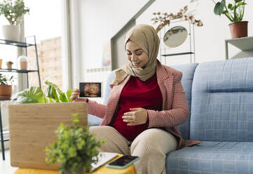 Schwangere Frau zeigt Ultraschallbild bei Videoanruf über Laptop im Wohnzimmer - JCCMF05065
