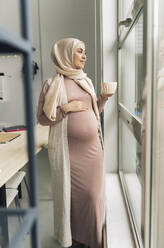 Nachdenkliche schwangere Frau, die durch das Fenster ihres Hauses schaut - JCCMF05113