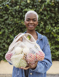 Smiling woman holding mesh bag with groceries - JCCMF05124